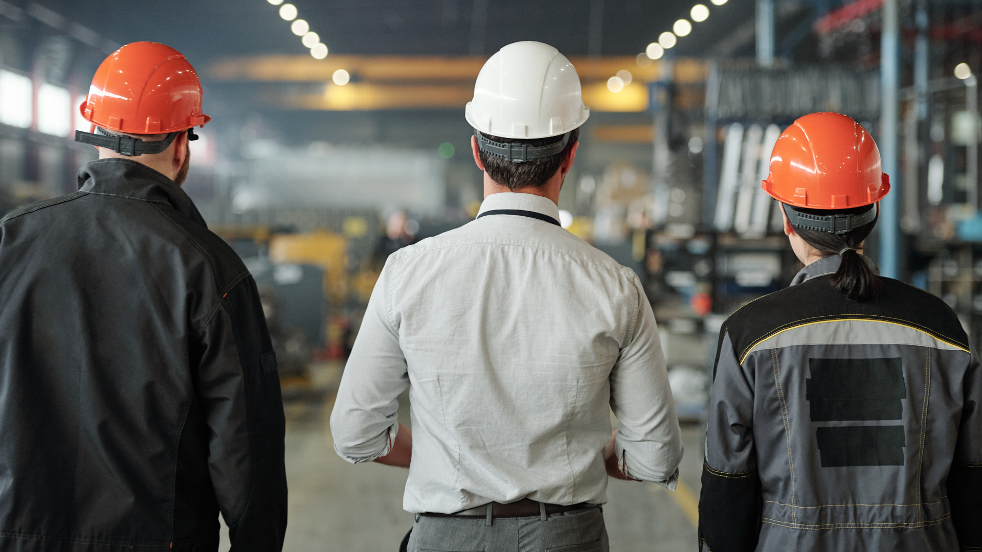 Three workers wearing safety helmets, including a white hard hat and orange hard hats, in an industrial environment.