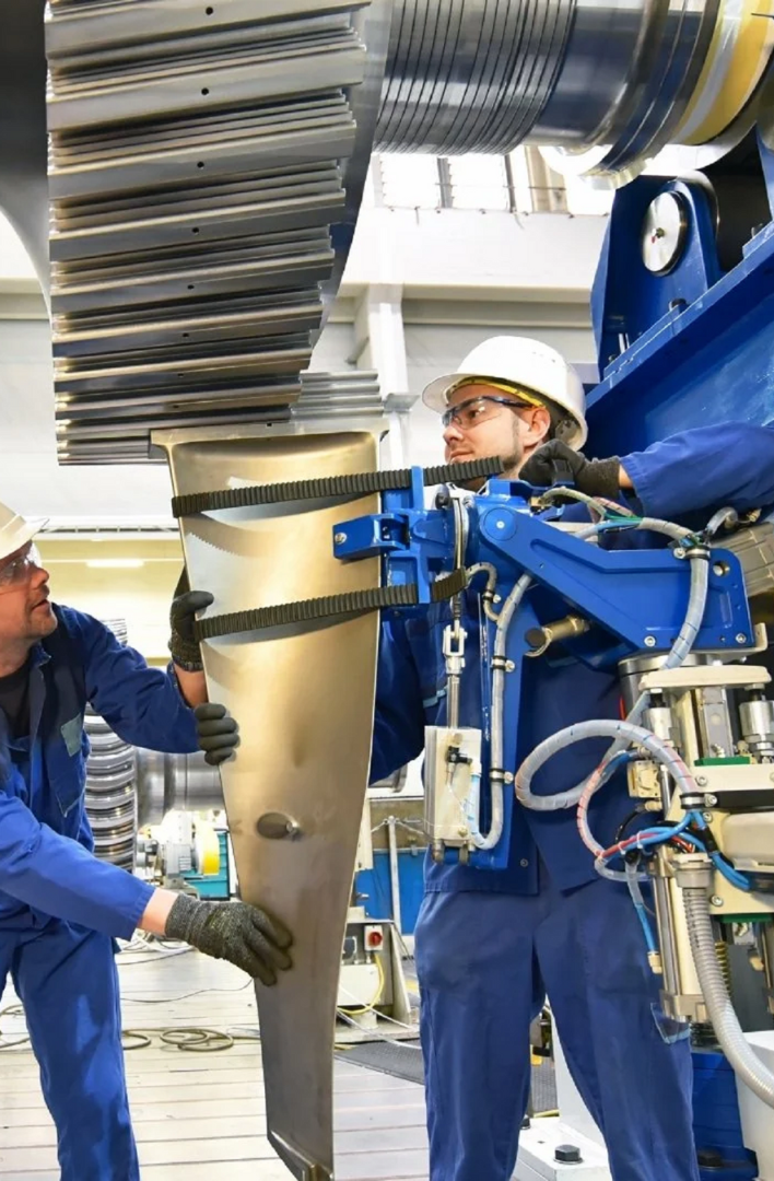 Two workers in blue uniforms and safety helmets assembling a large machinery component in a manufacturing facility.