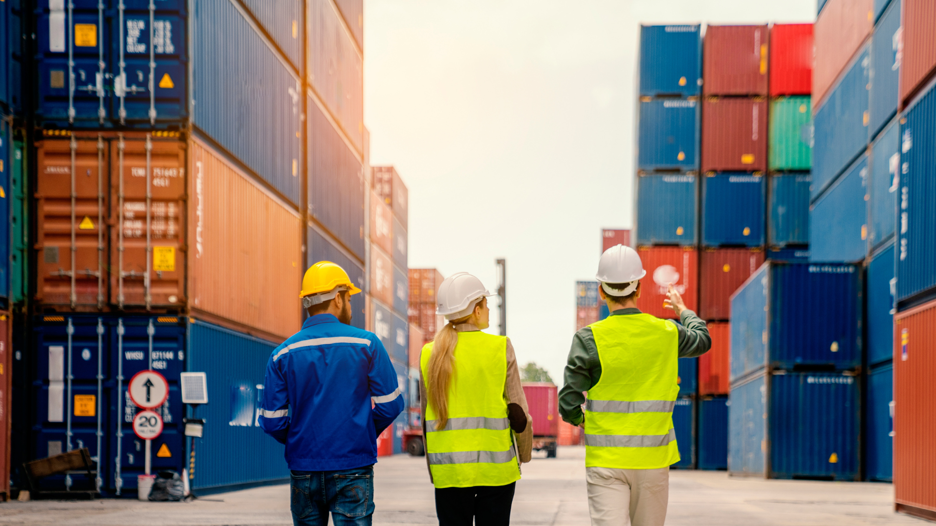 Three individuals in safety gear walking among shipping containers in a logistics yard.